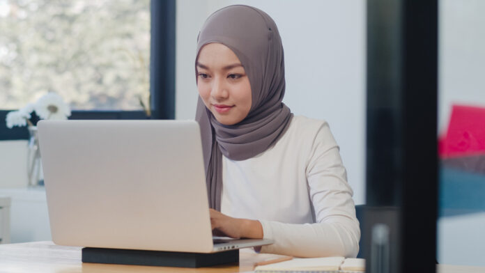 Asian Muslim lady in casual wear working on a laptop in a modern office setting, highlighting the importance of skill development and upskilling for success in the current job market